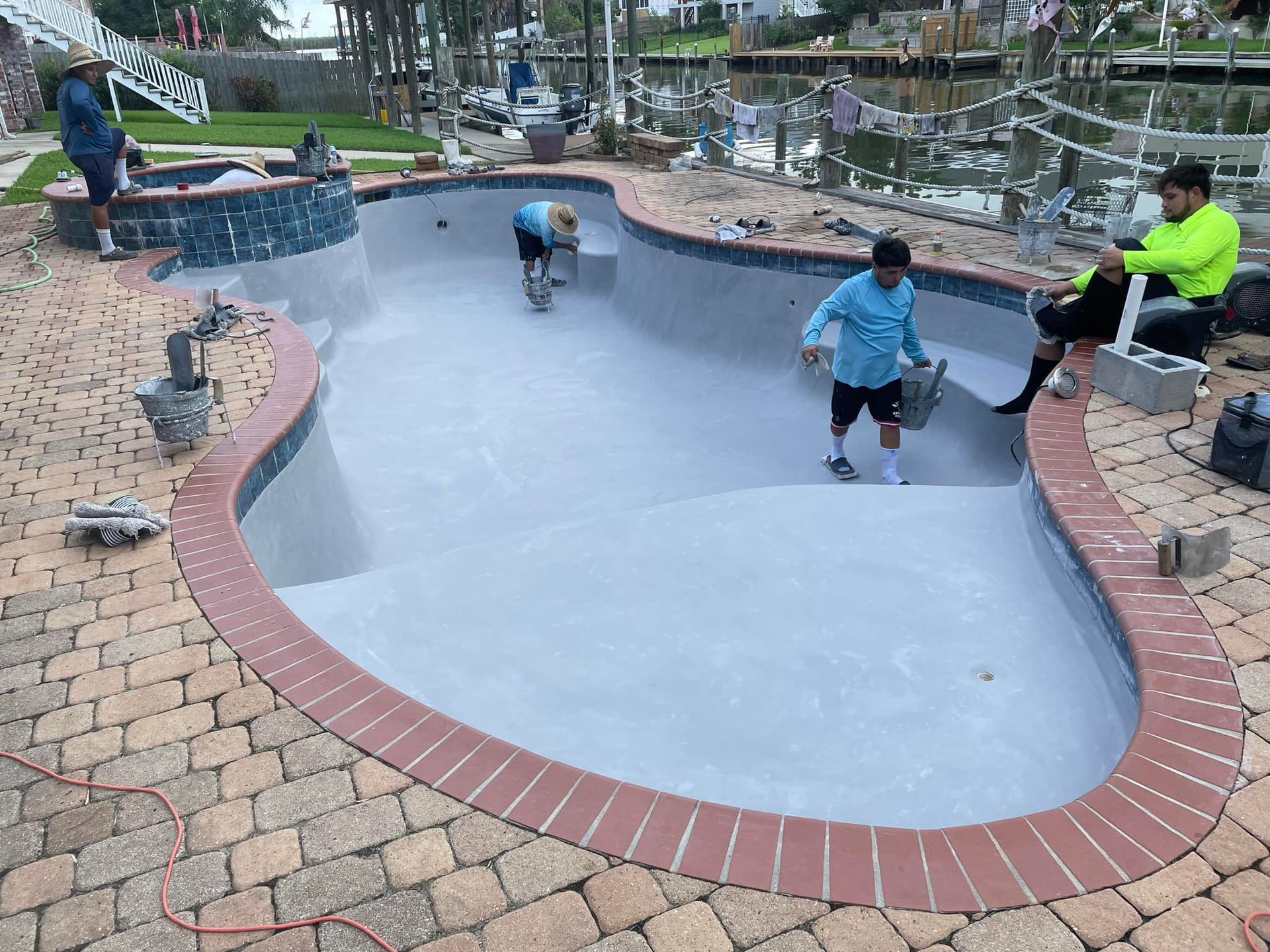People skating in a small empty concrete skate bowl beside a brick path and benches.