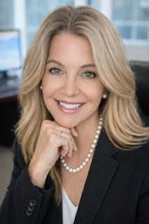 Woman smiling, wearing a black blazer over a red top. Dark, wavy hair. White background.