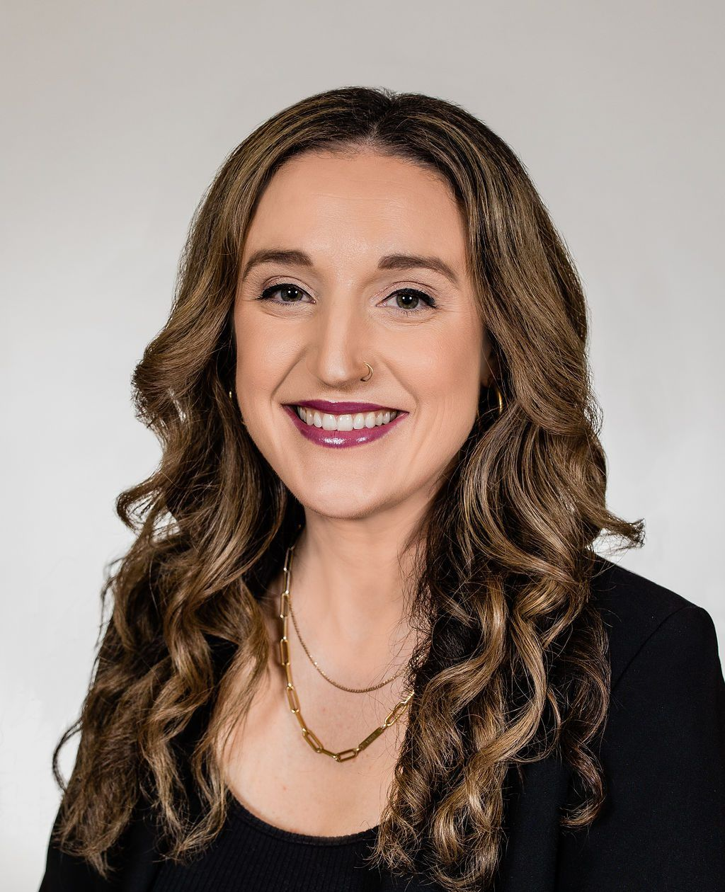 Woman with long wavy brown hair smiles, wearing a black blazer and gold necklace, against a white backdrop.