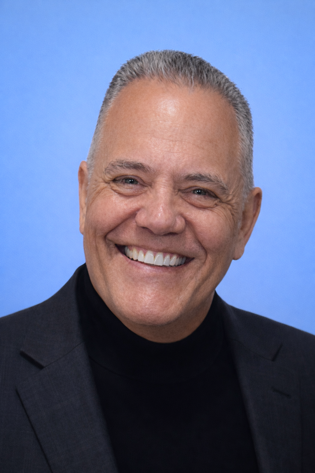 Woman smiling, wearing a black blazer over a red top. Dark, wavy hair. White background.