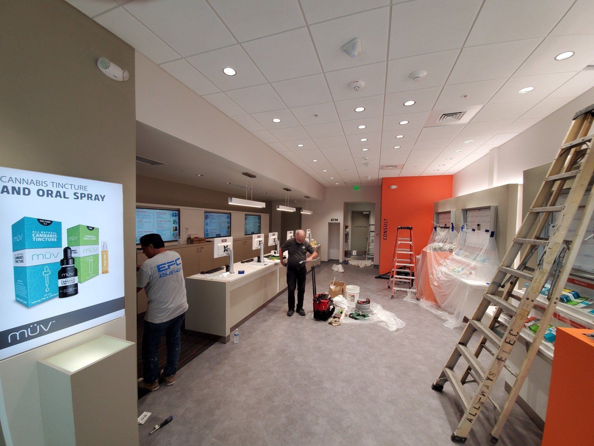 Interior of a store under construction, workers, ladders, display counters, product displays, and orange accent wall.