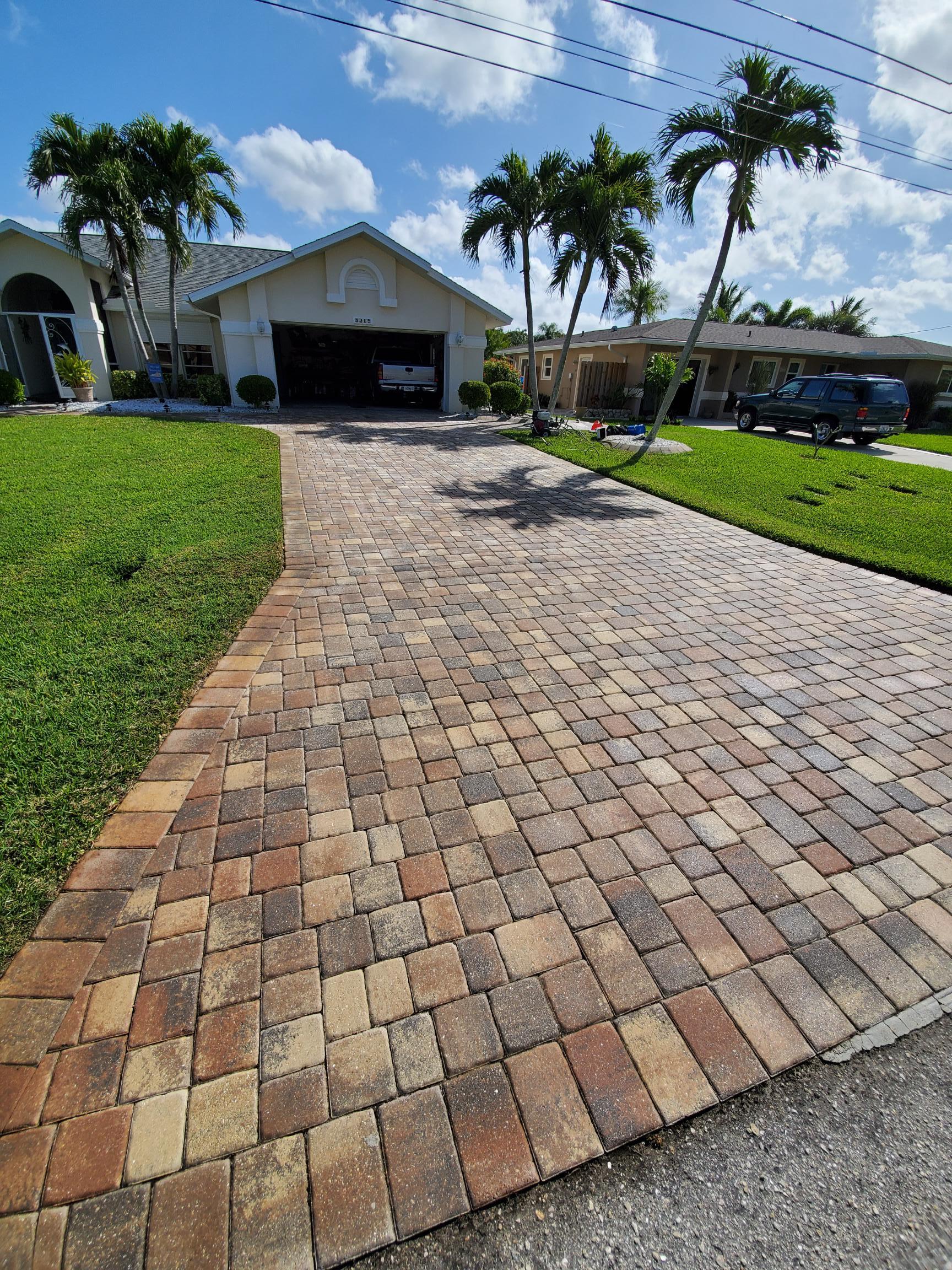 Brick paver driveway leading to a house with a garage, green grass, palm trees under a blue sky.