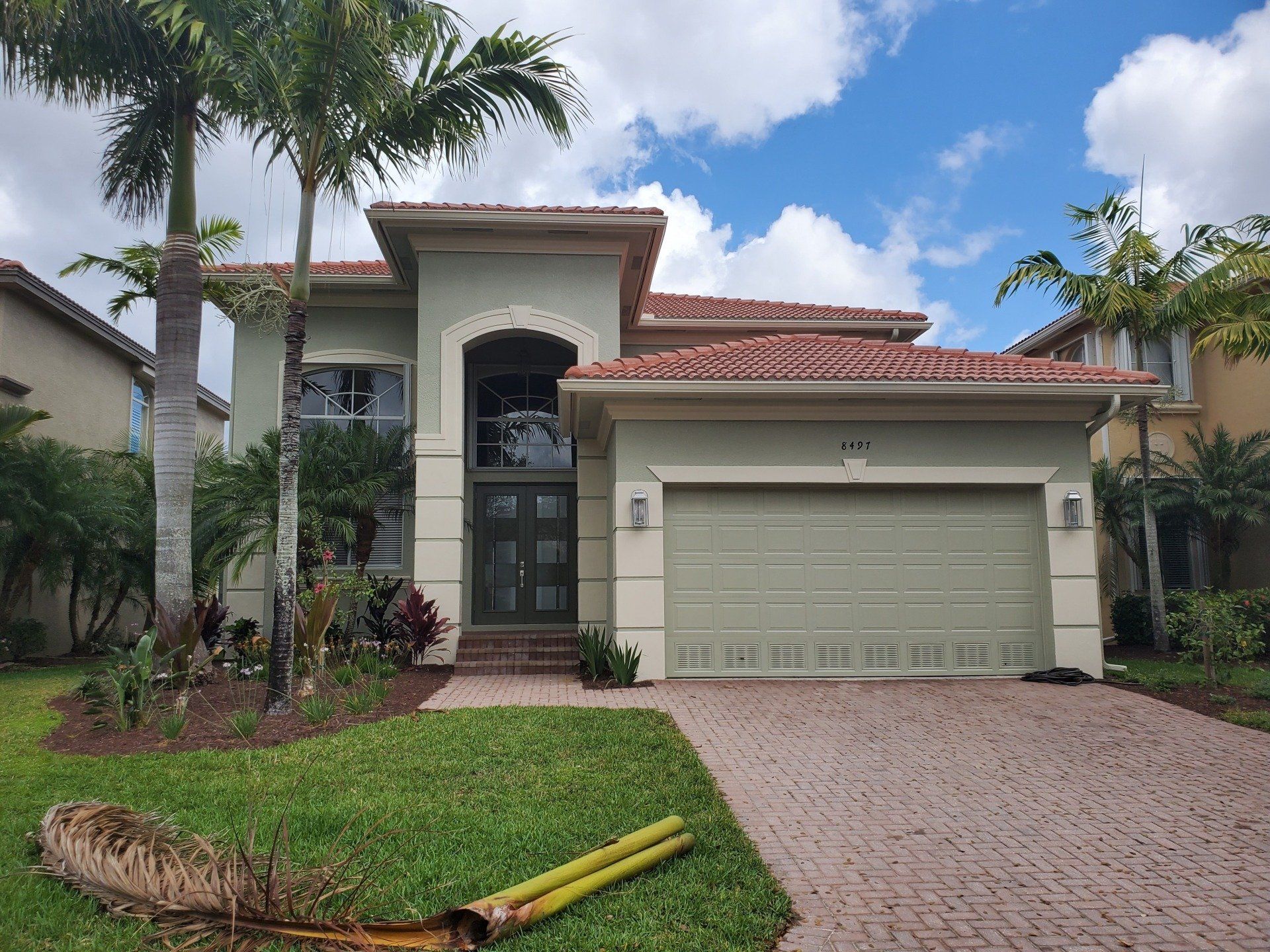 A two-story house with a green door and garage, palm trees, and a brick driveway.