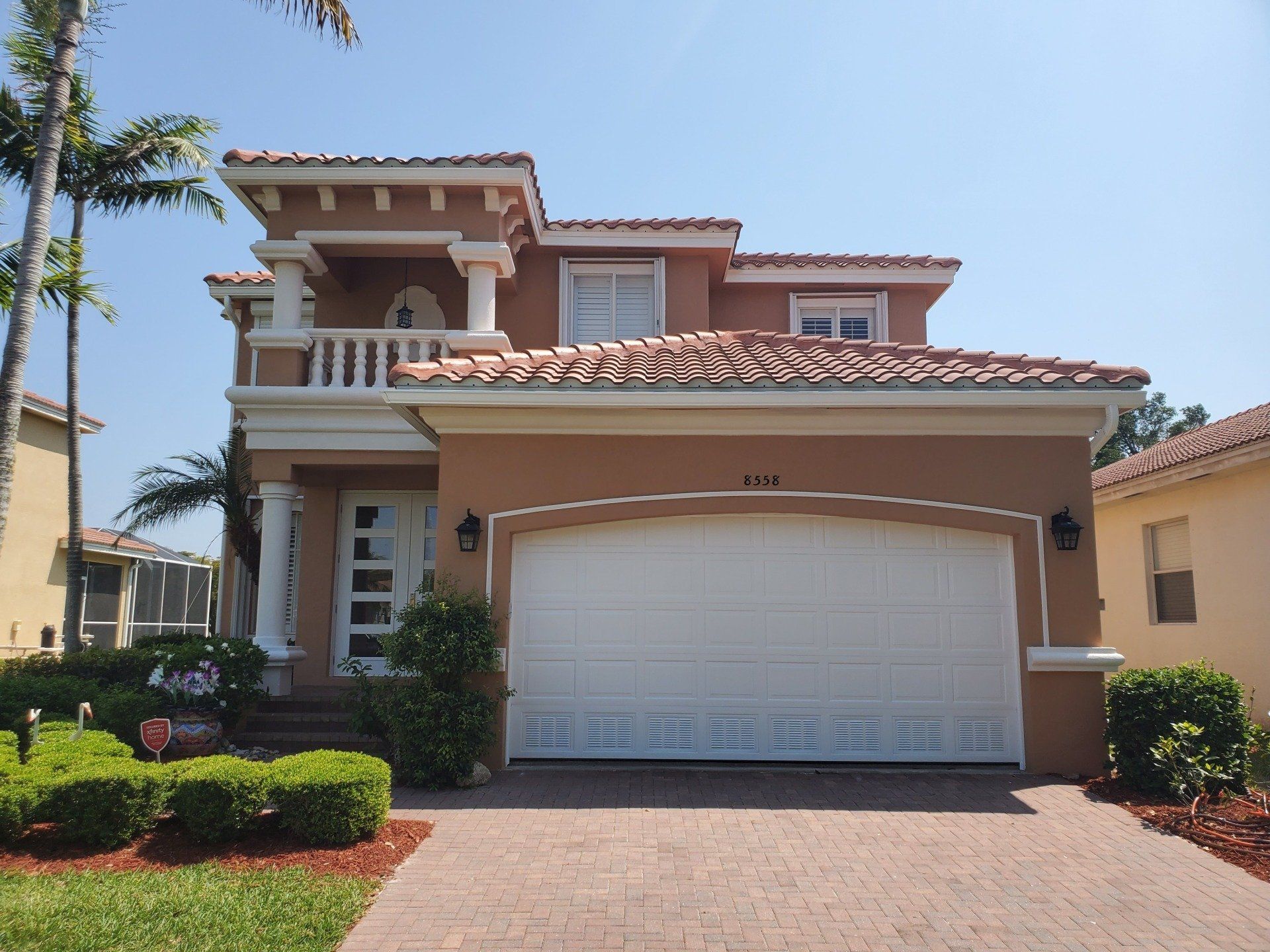 Two-story stucco house with a red tile roof, white garage door, and brick driveway.