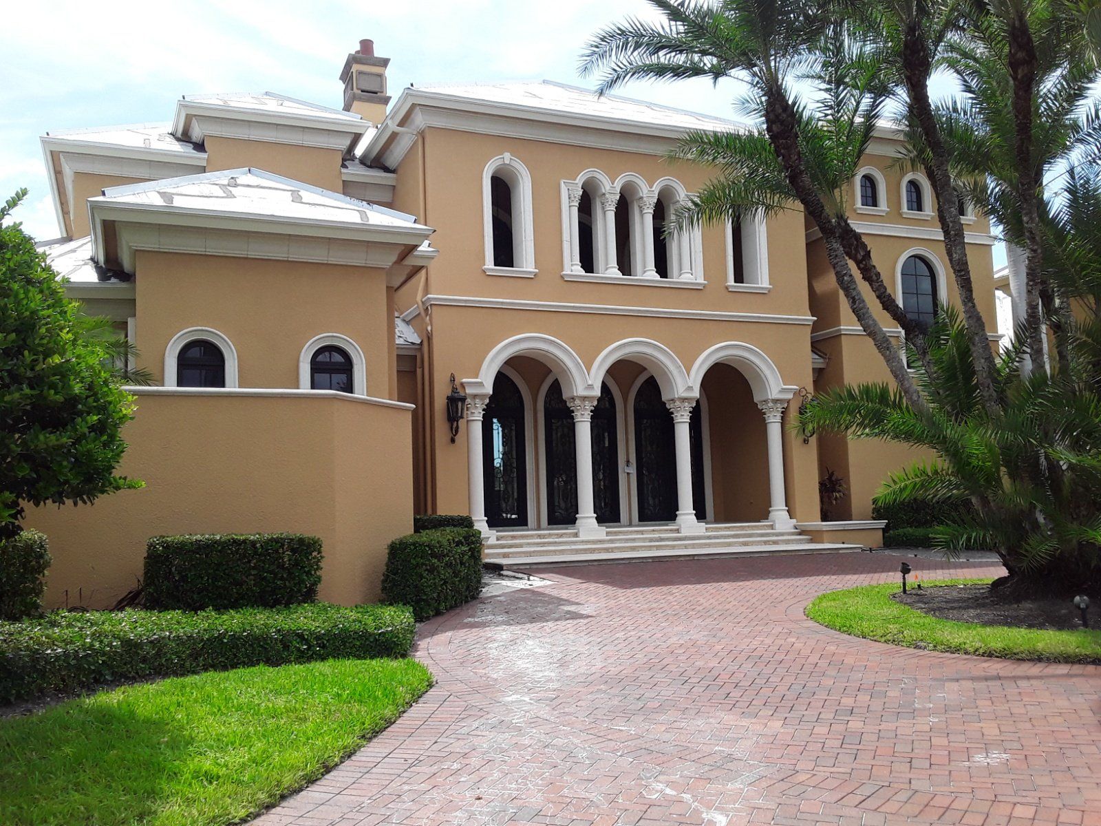 Tan mansion with white trim and arched entryway. Red brick driveway. Green grass and bushes.