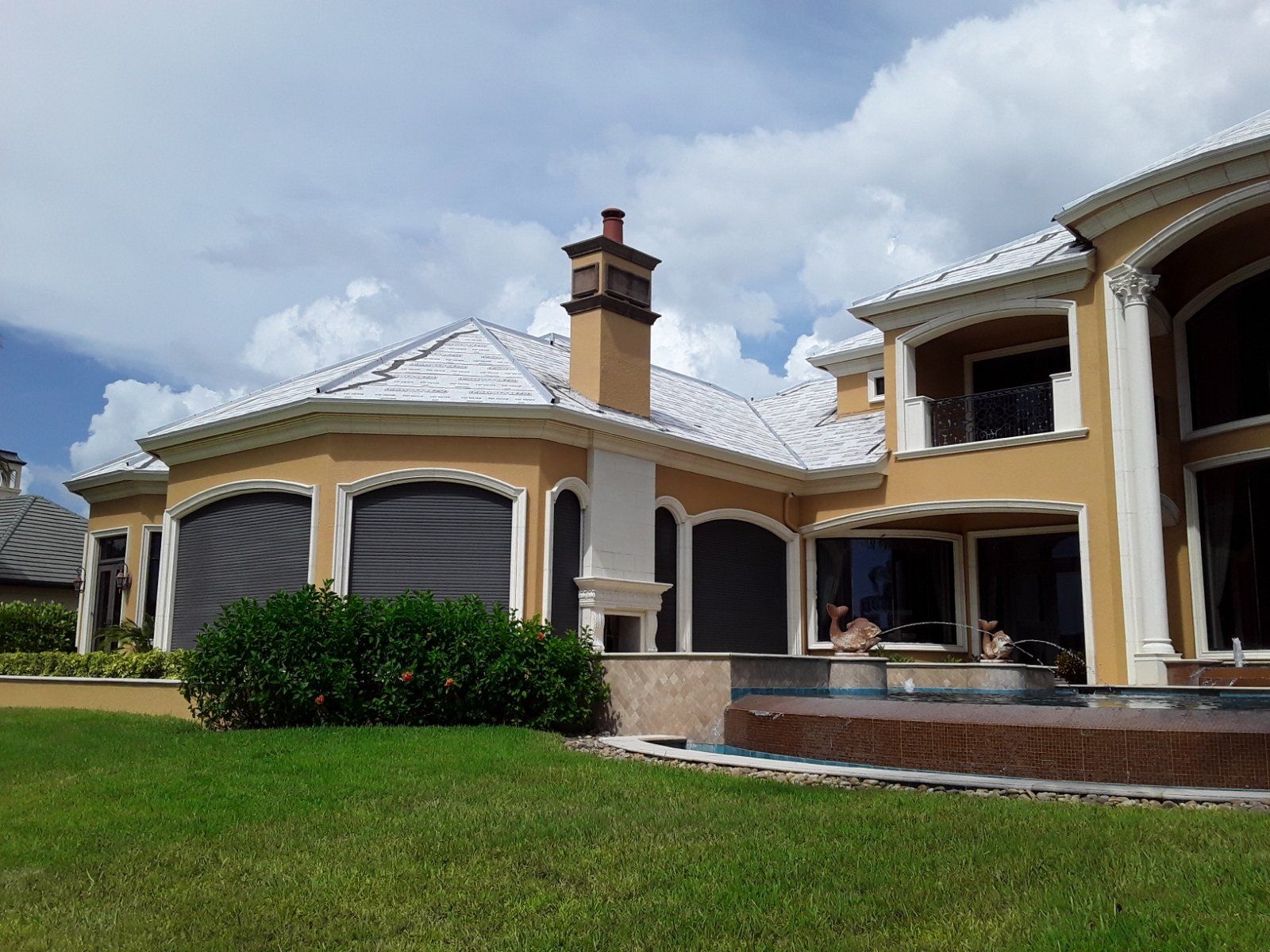 Large yellow mansion with white roof, chimney, and green lawn under blue sky with clouds.