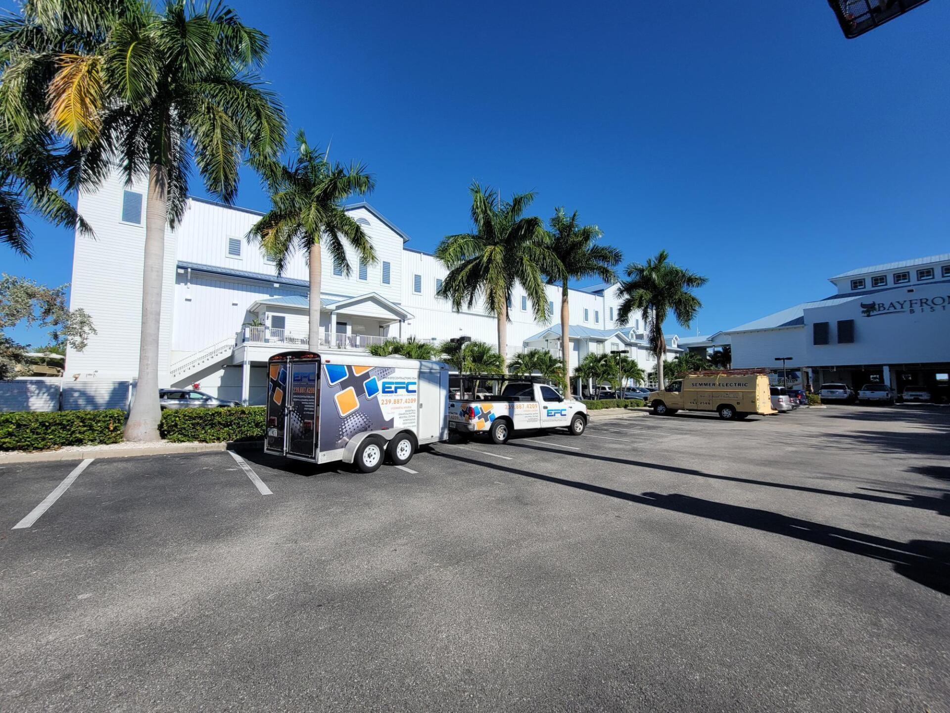 A parking lot with service vehicles in front of white buildings and palm trees on a sunny day.