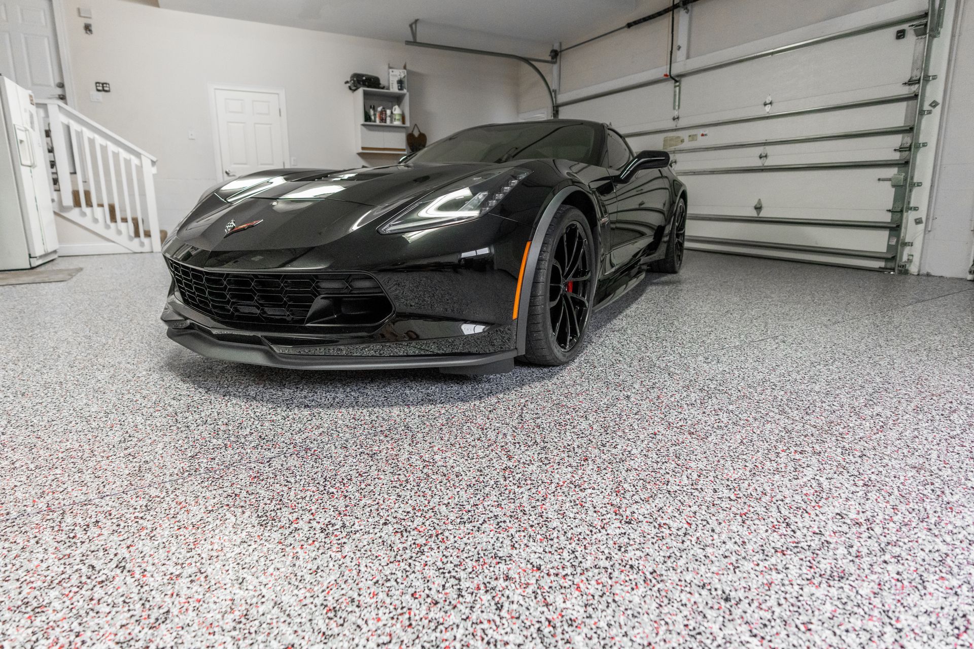 Black sports car parked in a garage with a speckled floor. Garage door is open.