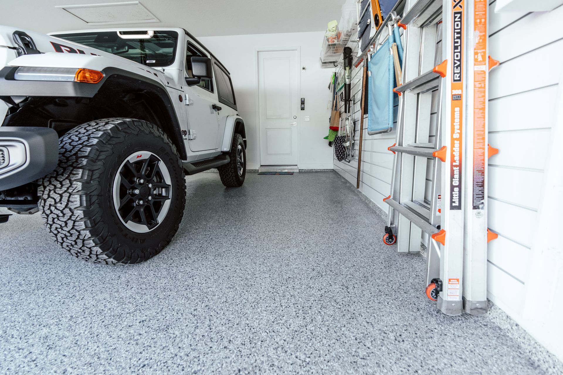 White Jeep parked in a garage with speckled gray flooring, ladder, and tools on the wall.