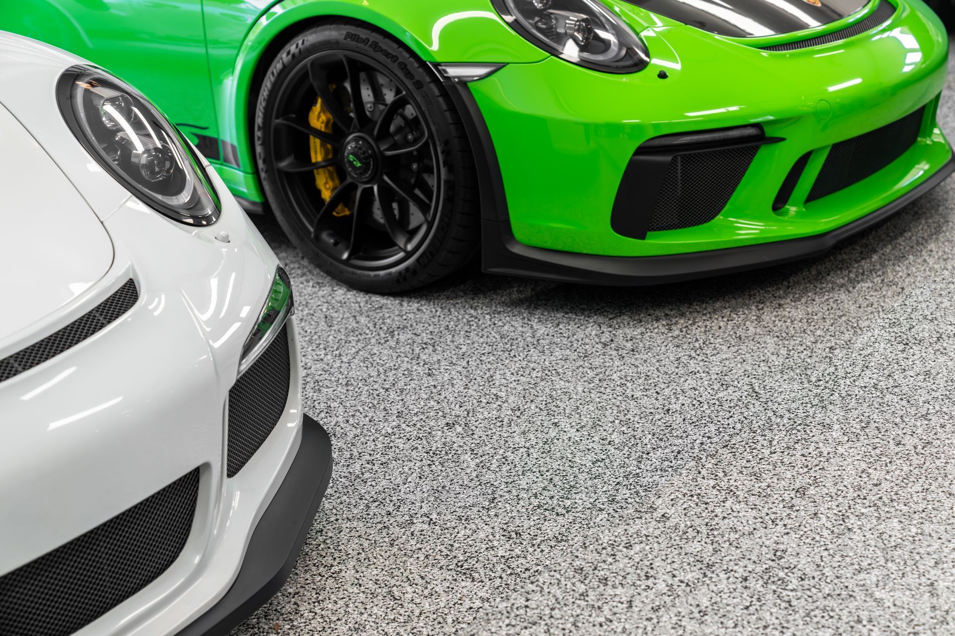 White and green Porsche sports cars parked on a speckled floor, close-up shot of front ends.