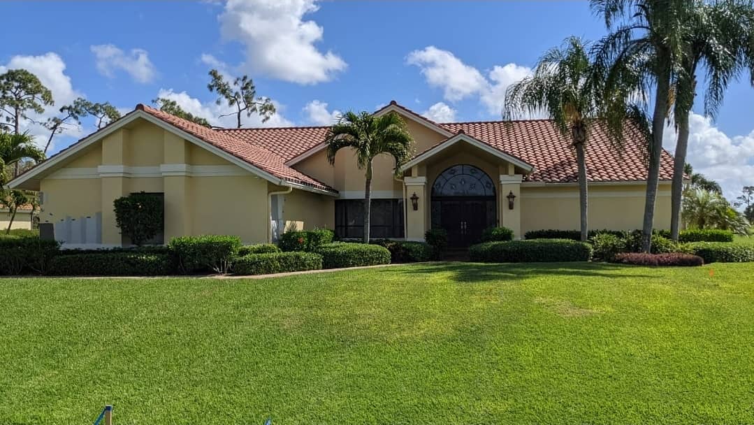 Yellow stucco house with red tile roof, palm trees, and green lawn under blue sky.