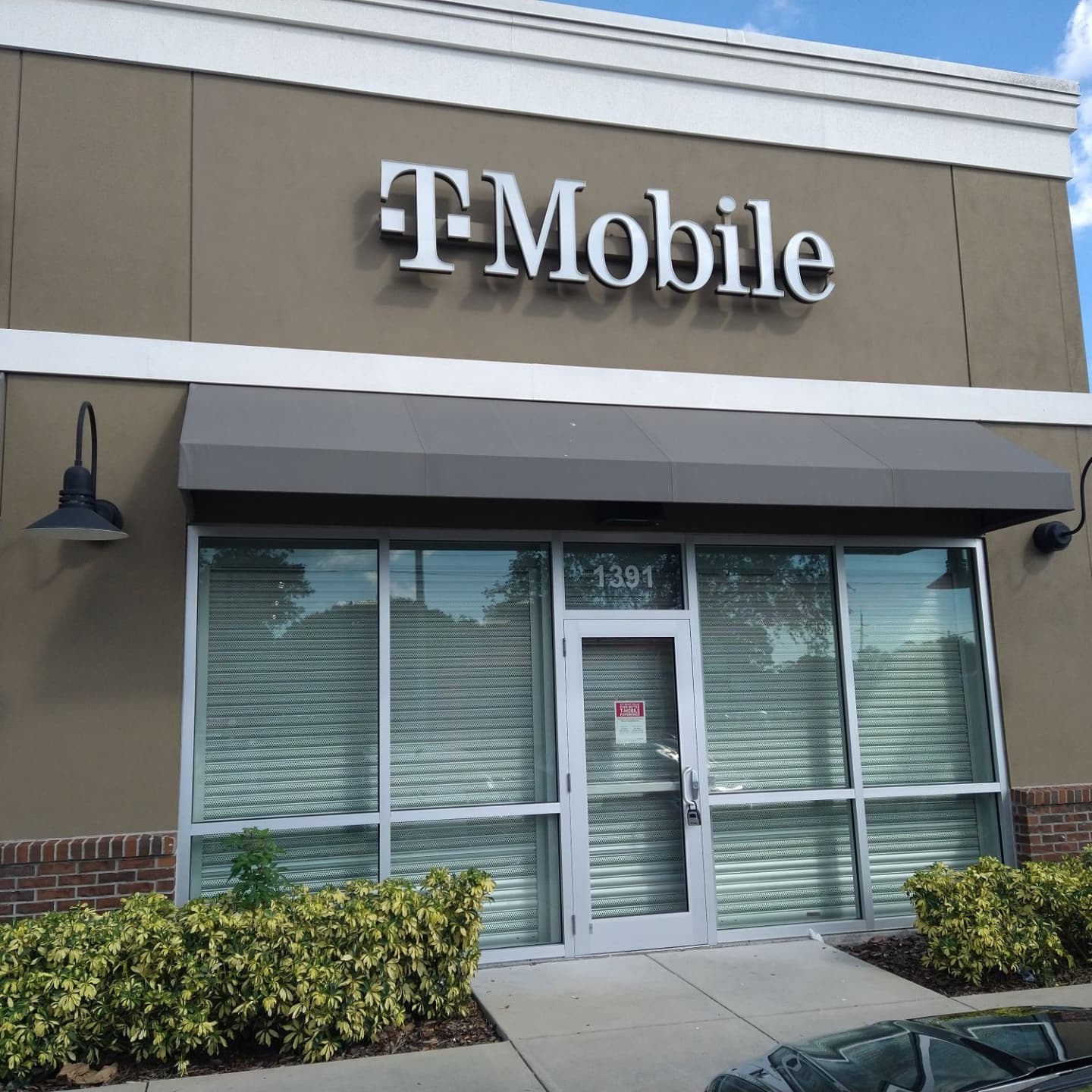 T-Mobile store front with gray awning, sign, and windows; bushes in front.