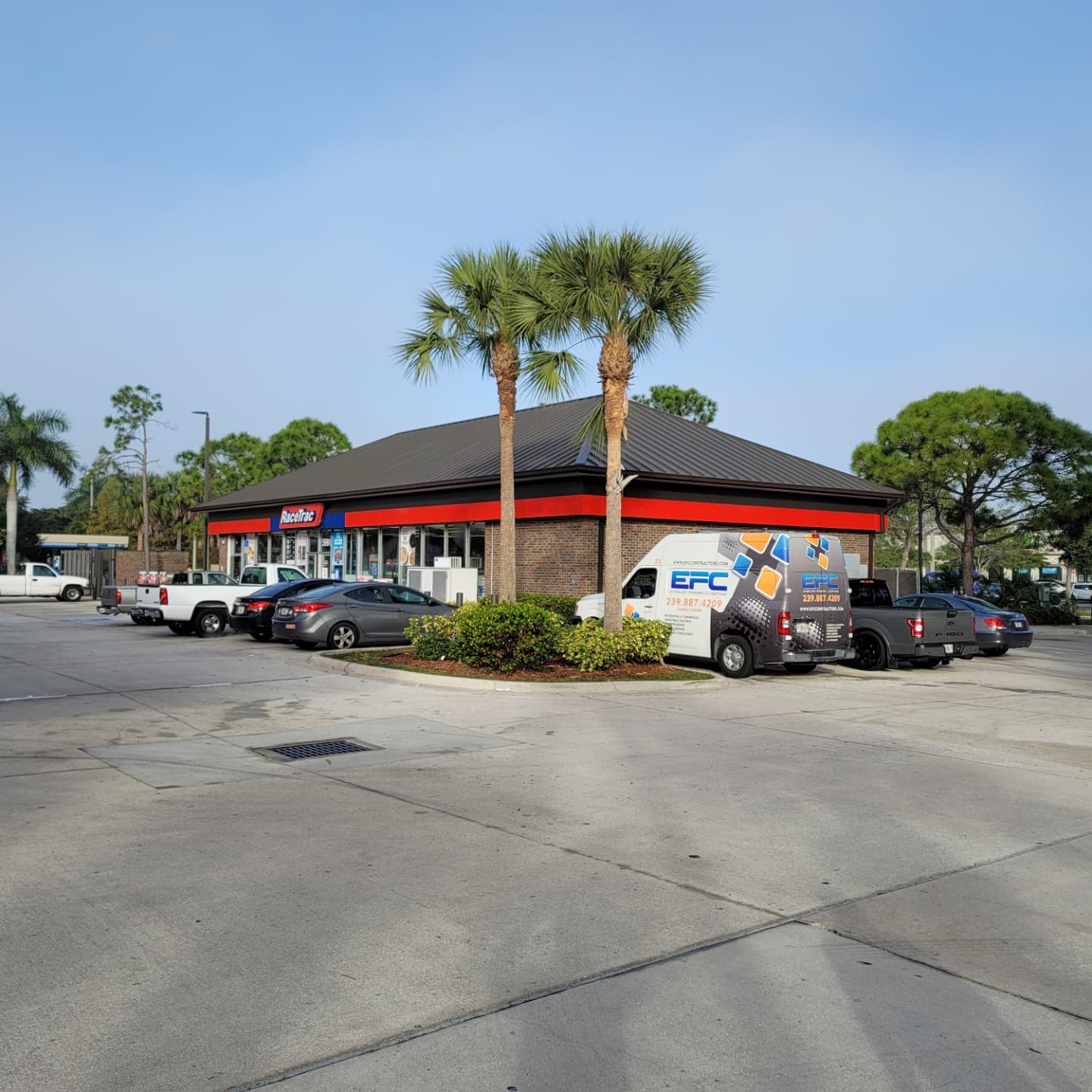 A convenience store with a red and black facade. Palm trees stand out front, and vehicles are parked nearby.