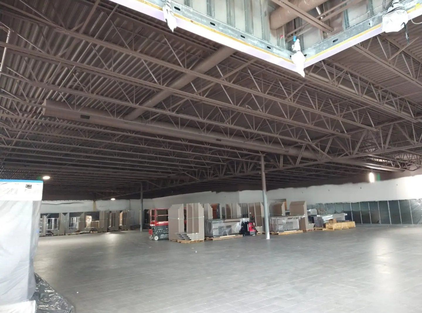 Interior of a large, empty warehouse with exposed steel beams and concrete floor. Boxes and a forklift are visible.