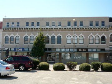 Beige three-story building with arched windows, parking lot, vehicles, and business signage.