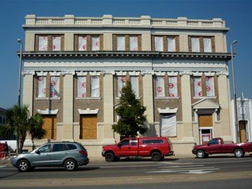 Beige, two-story building with columns; windows boarded up, parked cars on street.