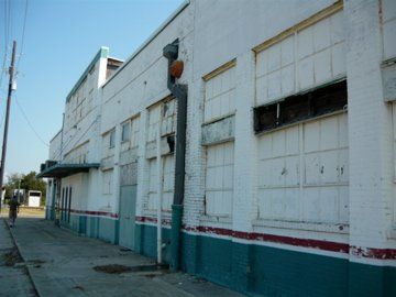 Dilapidated white brick building with broken windows and teal accents; person walks on sidewalk.