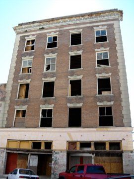 Abandoned multi-story brick building with boarded-up windows and broken windows. Cars parked in front.