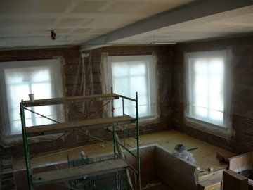 Interior room under construction with three windows, scaffolding, and exposed brick walls.