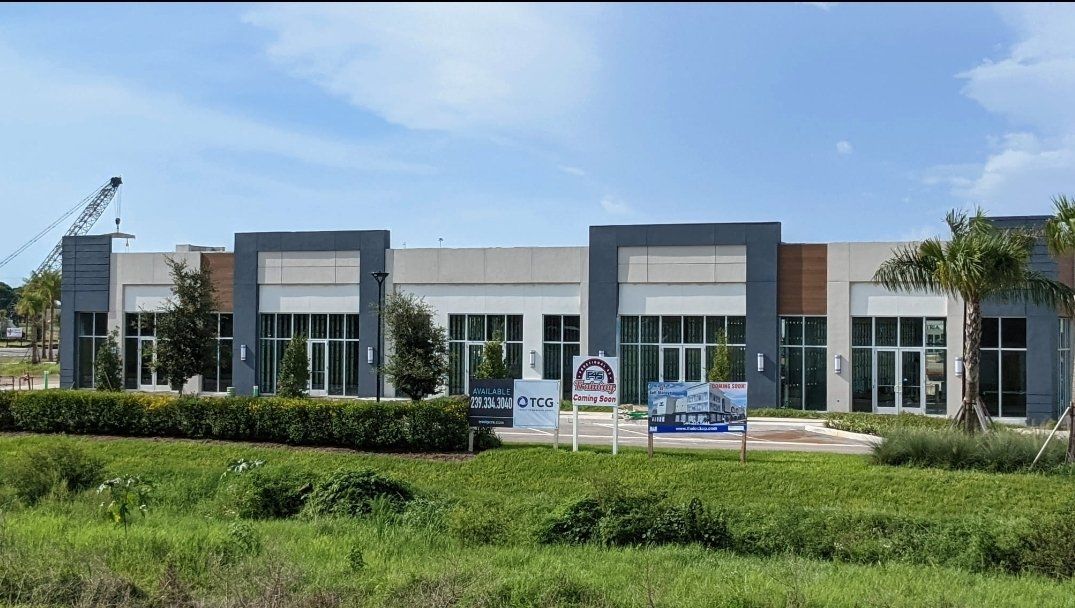 Modern commercial building with gray and white facade, glass windows, and landscaping under a blue sky.