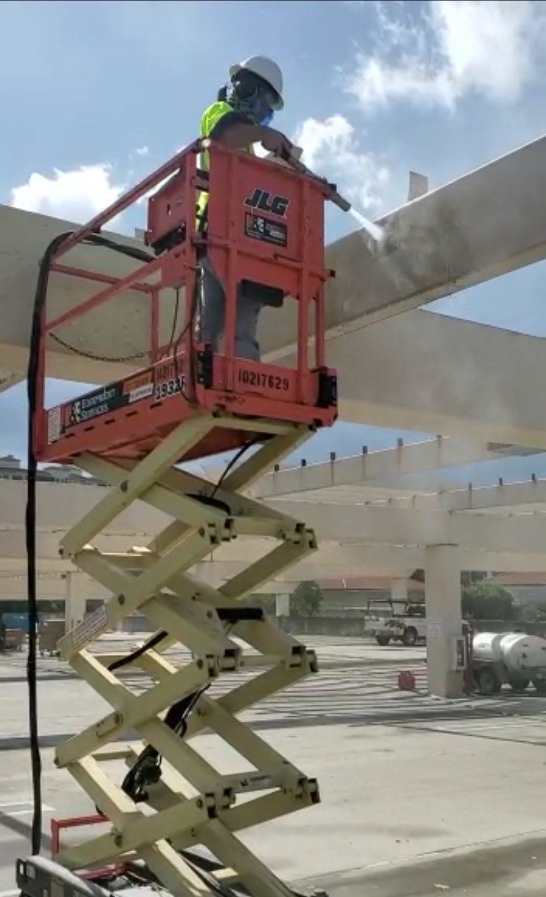 Construction worker spraying concrete beam from an elevated lift. Bright day, safety gear.