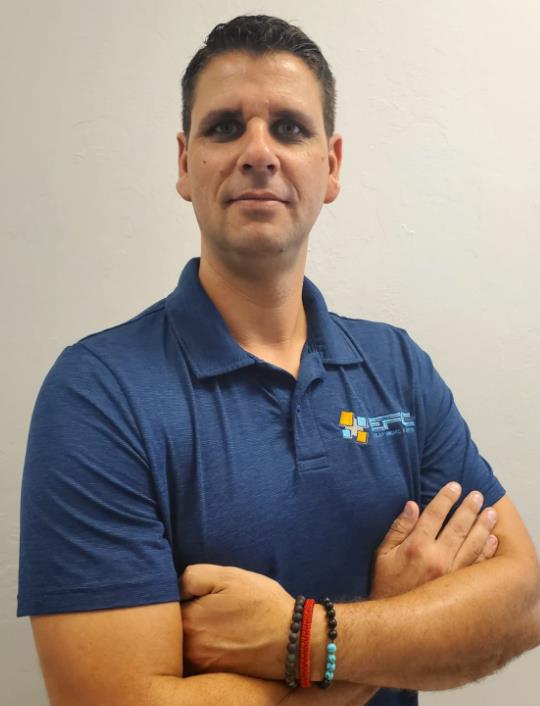 Man with arms crossed, wearing blue polo shirt, smiling, standing in front of a white wall.