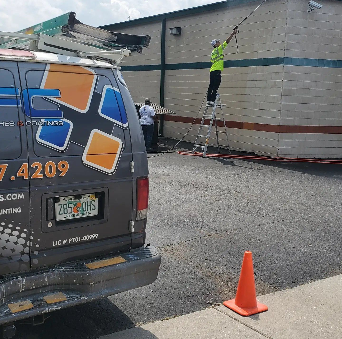 A worker on a ladder washing a building's exterior next to a work van. Orange traffic cone on the ground.