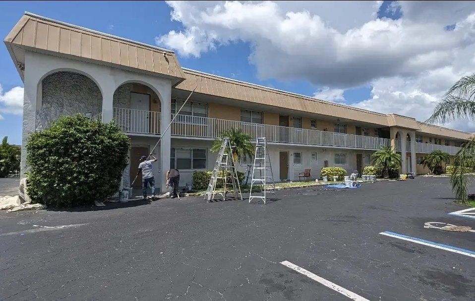 Two-story motel with tan roof and facade. Two workers on ladders. Parking lot in front, blue sky overhead.