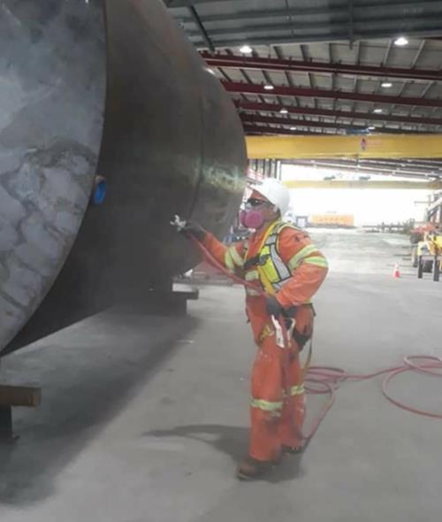 Worker in safety gear spraying paint on a large metal structure inside a warehouse.