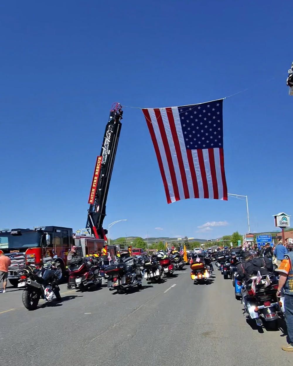 An American flag is suspended from a firetruck ladder over a road filled with motorcycles and riders