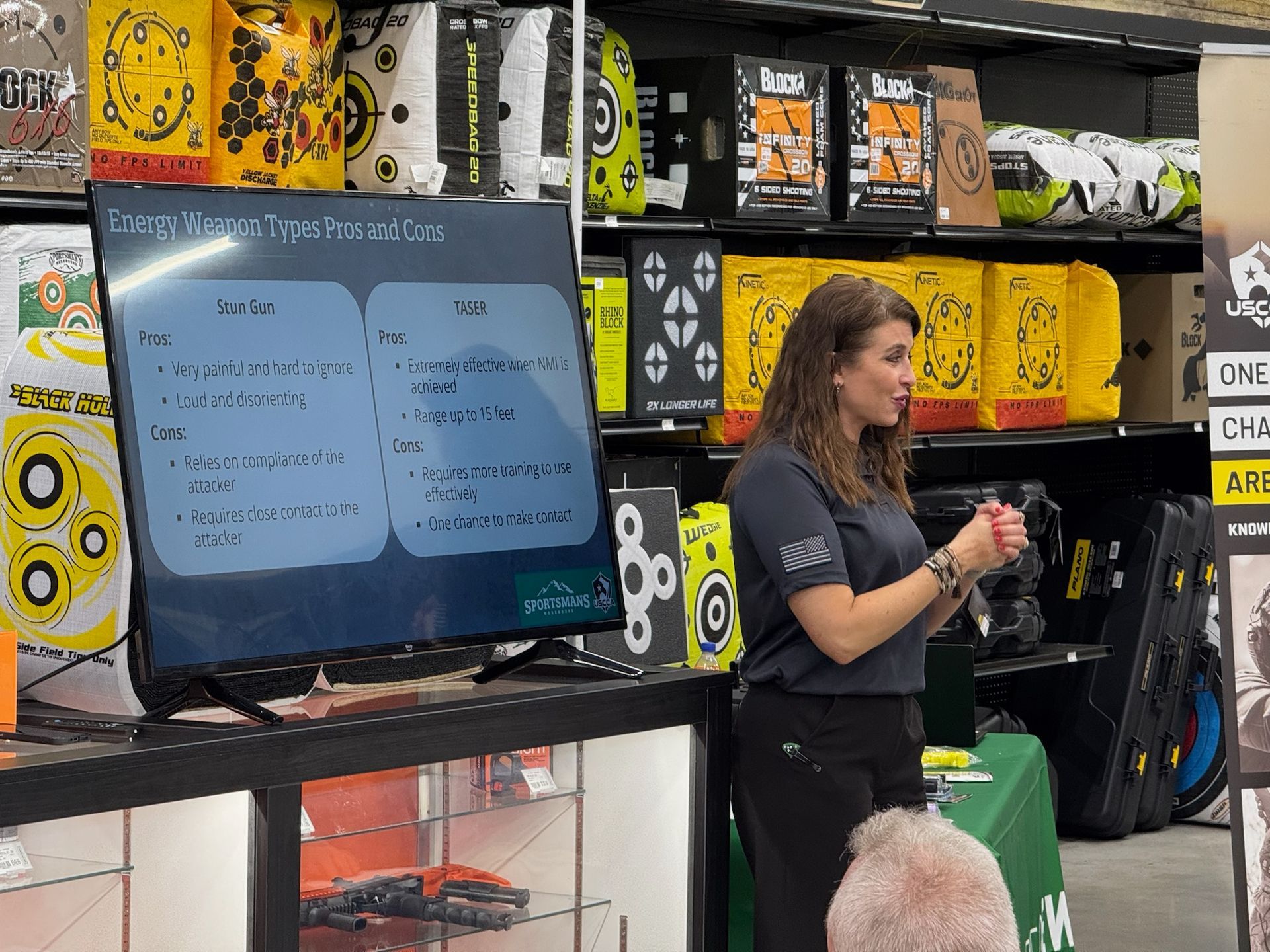 A woman wearing earmuffs and eye protection aims a handgun at a paper target inside an indoor shooting range booth.