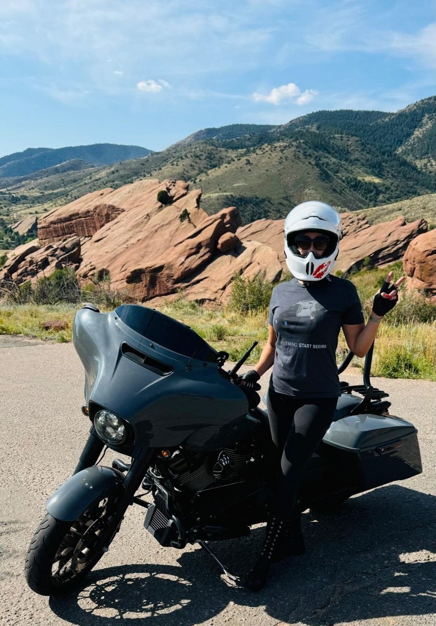 Amanda Hardin on a motorcycle, holding a rifle outside a shop with glass doors.