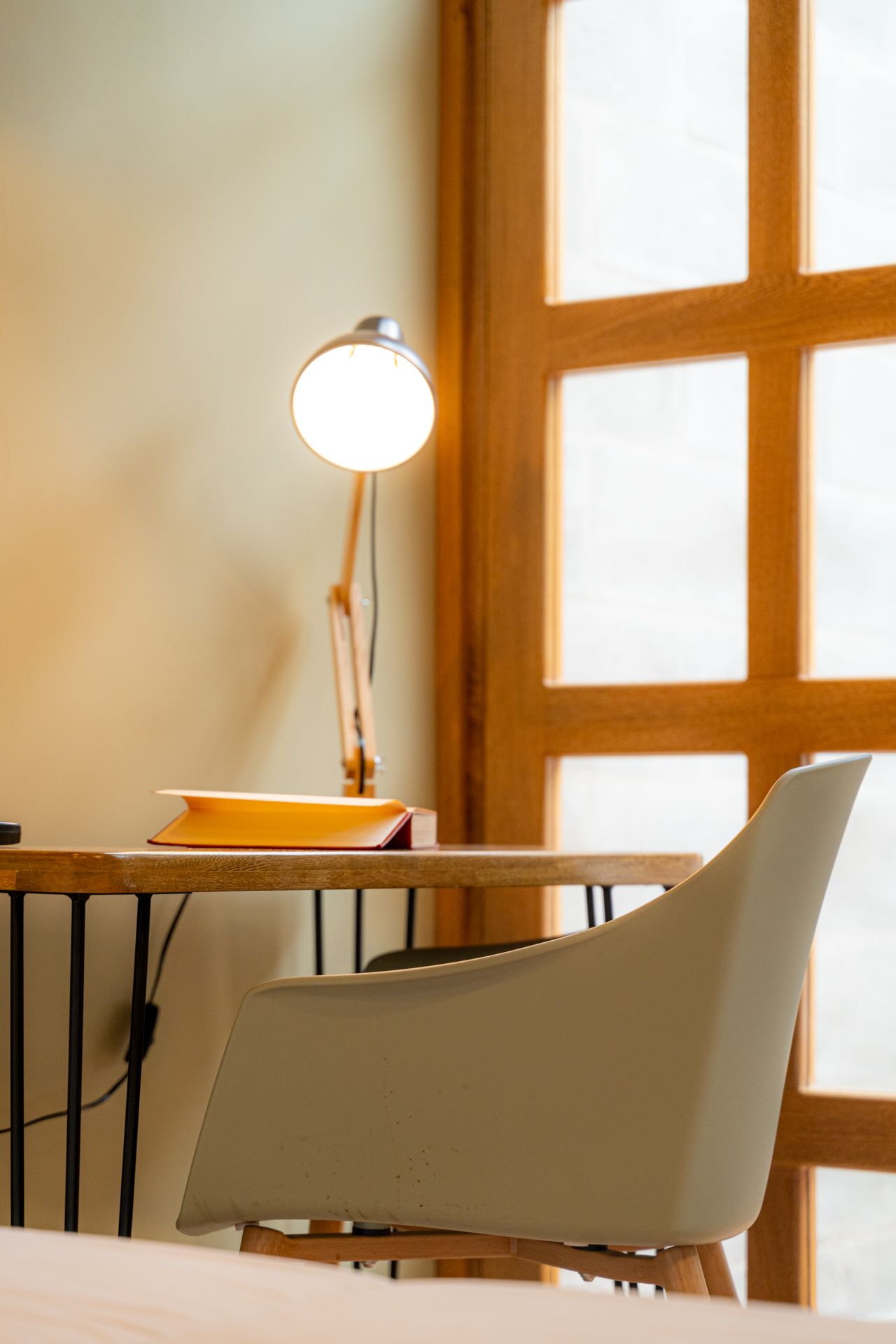 Escritorio con lámpara y libro, silla blanca, ventana de madera y pared beige claro.