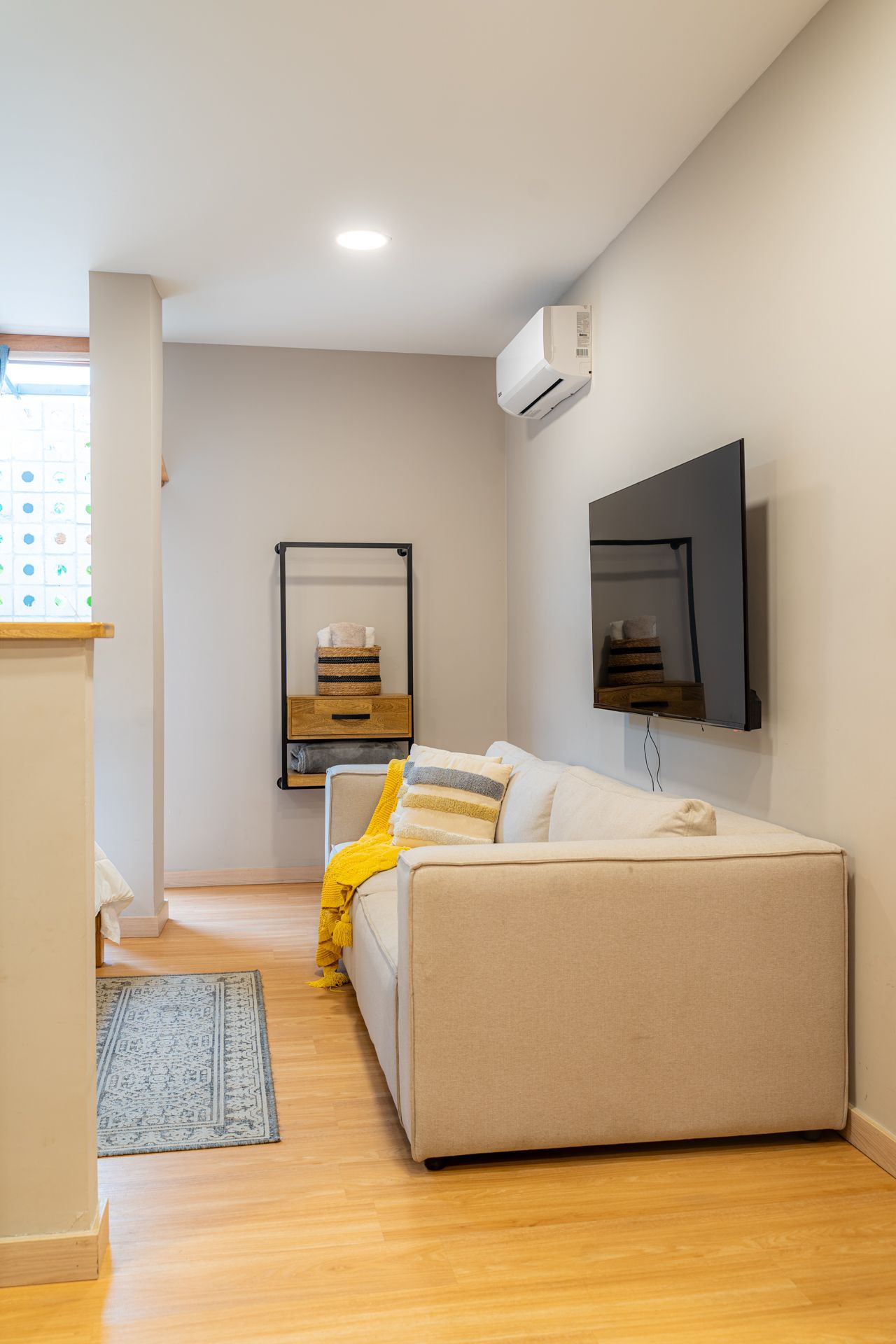 Pequeña sala de estar con sofá beige, TV, aire acondicionado y pisos de madera.