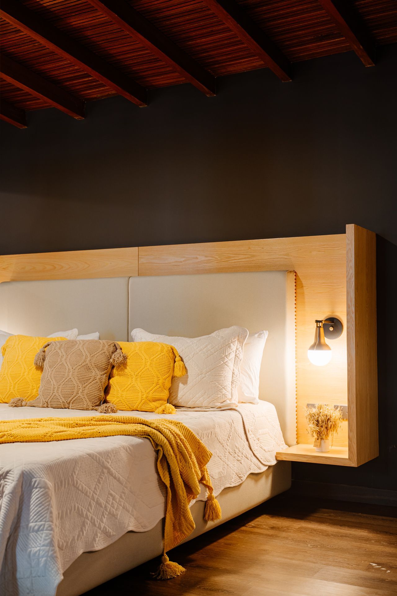 Dormitorio con cabecero de madera, almohadas y manta amarillas, aplique iluminado y pared oscura.