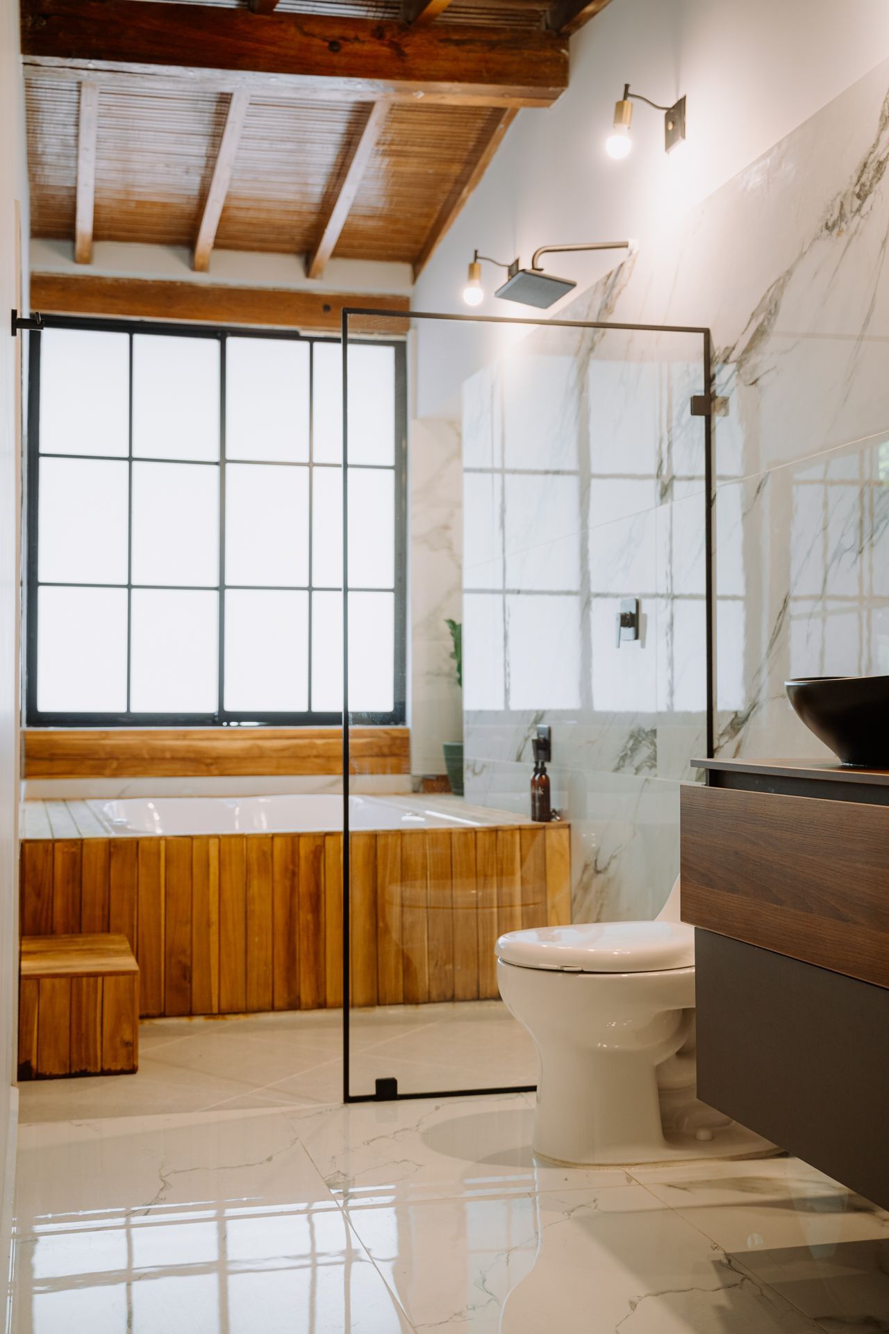 Baño moderno con detalles en madera, ventana grande, ducha de cristal, inodoro blanco, pisos de mármol.