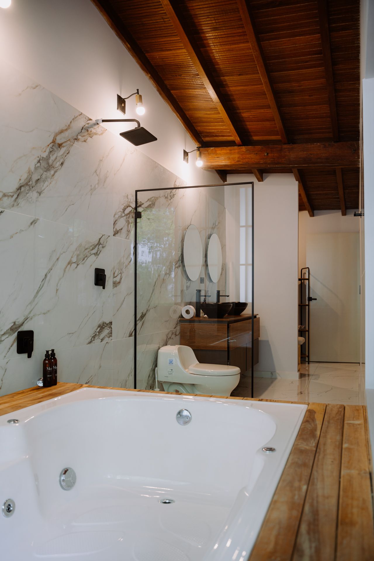 Baño con jacuzzi, pared decorativa de mármol, ducha de vidrio, techo de madera y tocador.