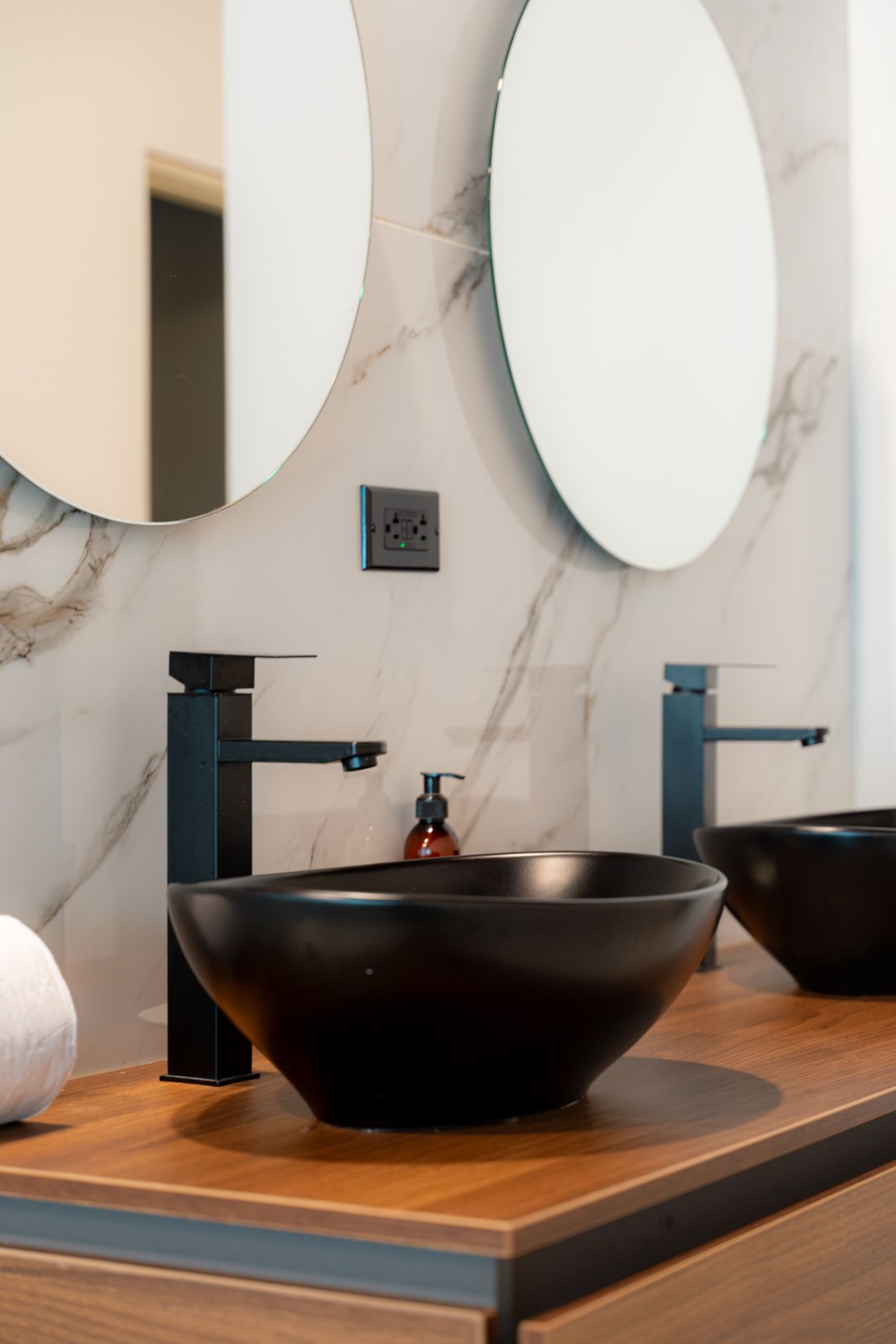 Baño moderno con lavabos de color negro, grifos negros, espejos redondos y encimera de madera.