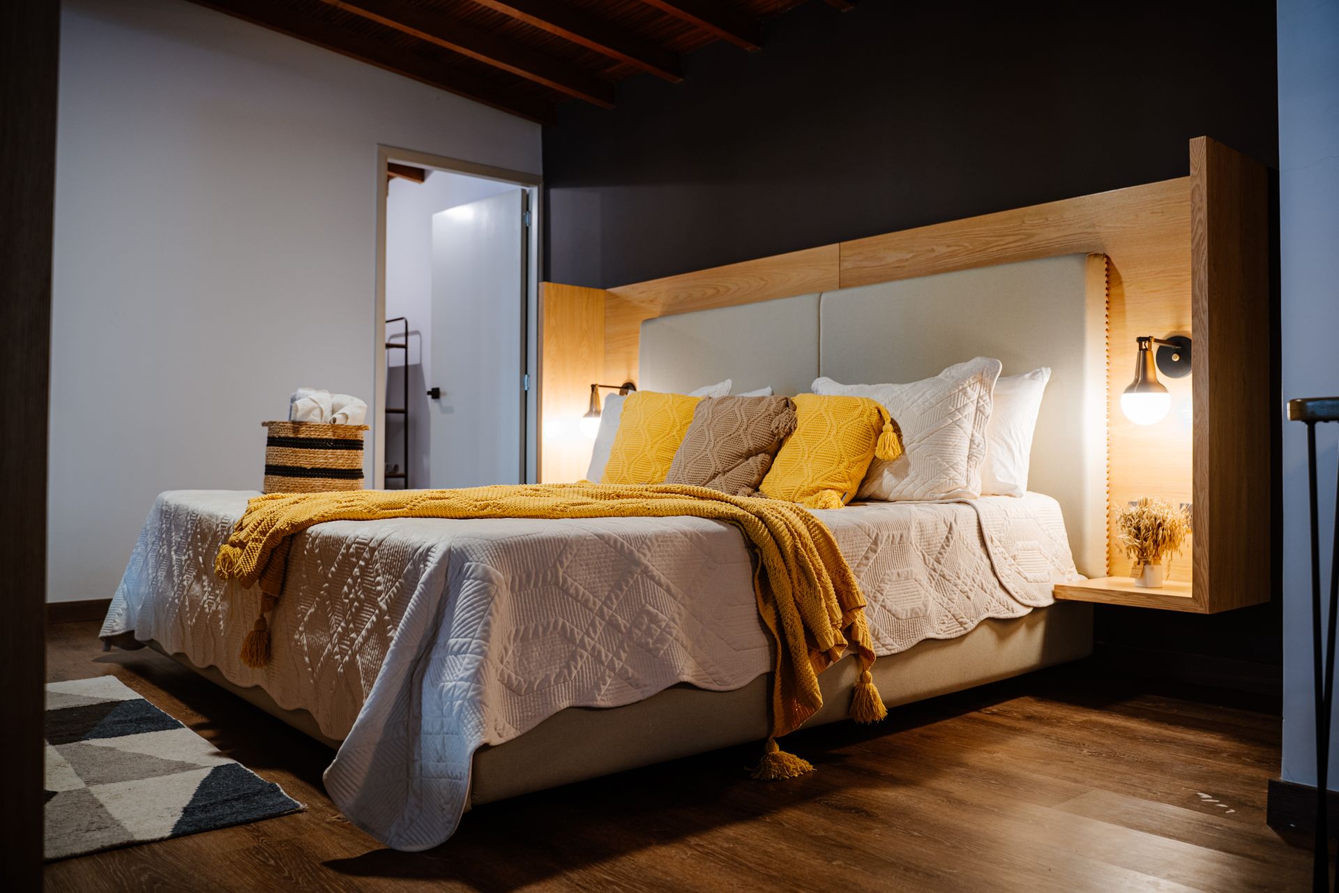 Dormitorio con cama, almohadas y manta amarilla, junto a una pequeña mesita de noche y un baño parcialmente visible.