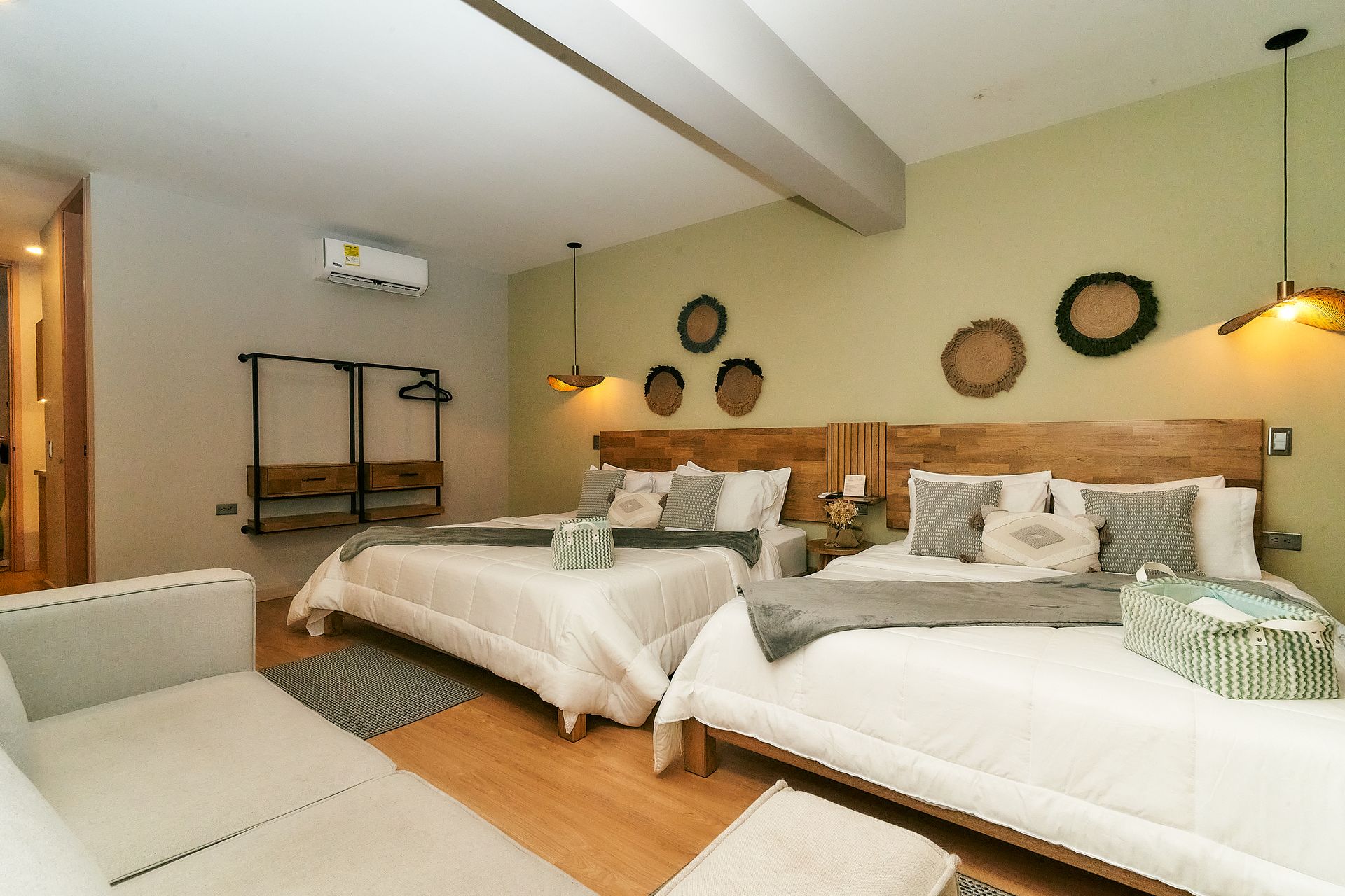 Habitación de hotel con dos camas, paredes verde claro, cabeceros de madera y un sofá blanco.