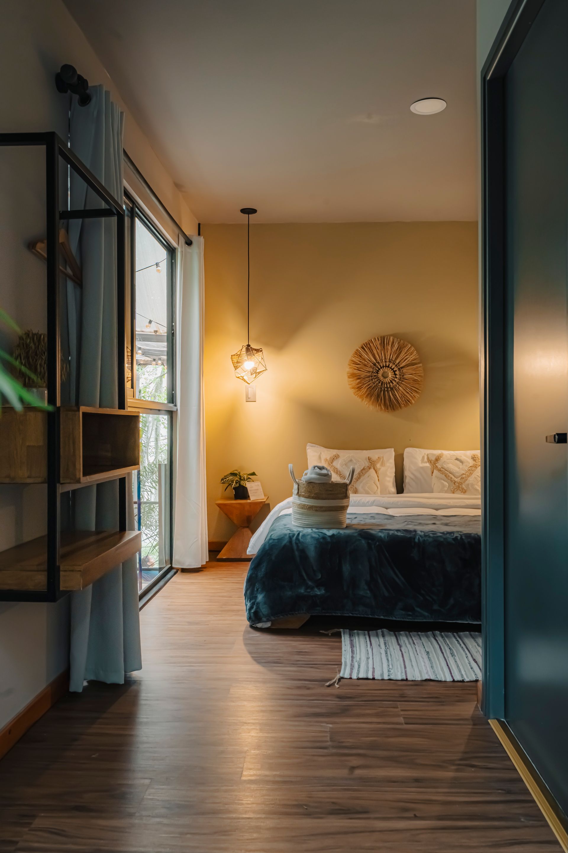 Interior de dormitorio con piso de madera, cama, ventana abierta, estante de almacenamiento y pared amarilla.