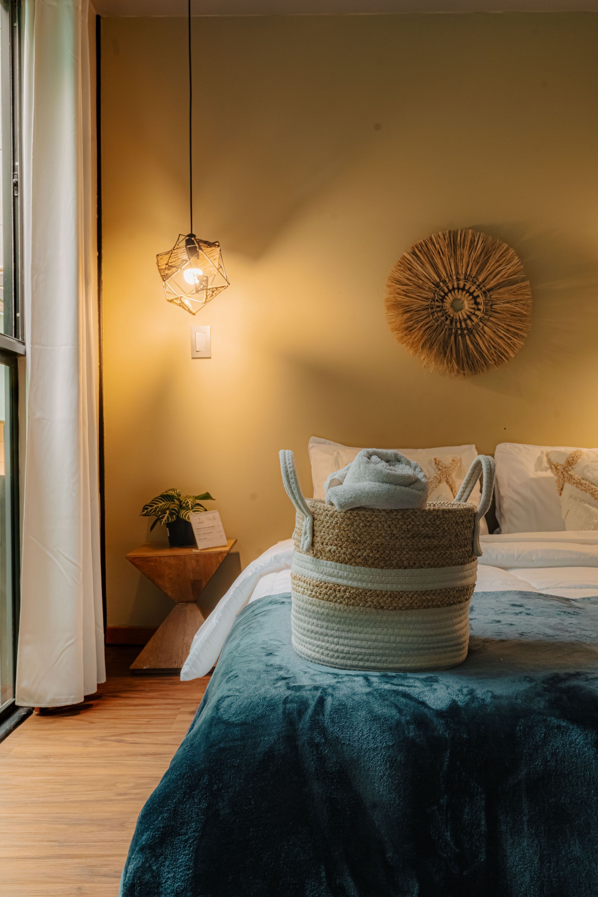 Dormitorio con manta azul, canasta tejida, lámpara colgante y arte de pared decorativo.