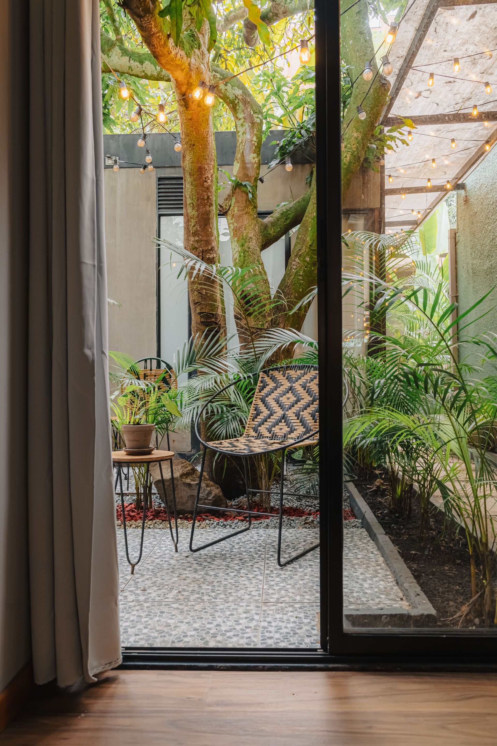 Vista a través de la puerta corrediza de vidrio del patio al aire libre con silla, árbol y luces de cadena.
