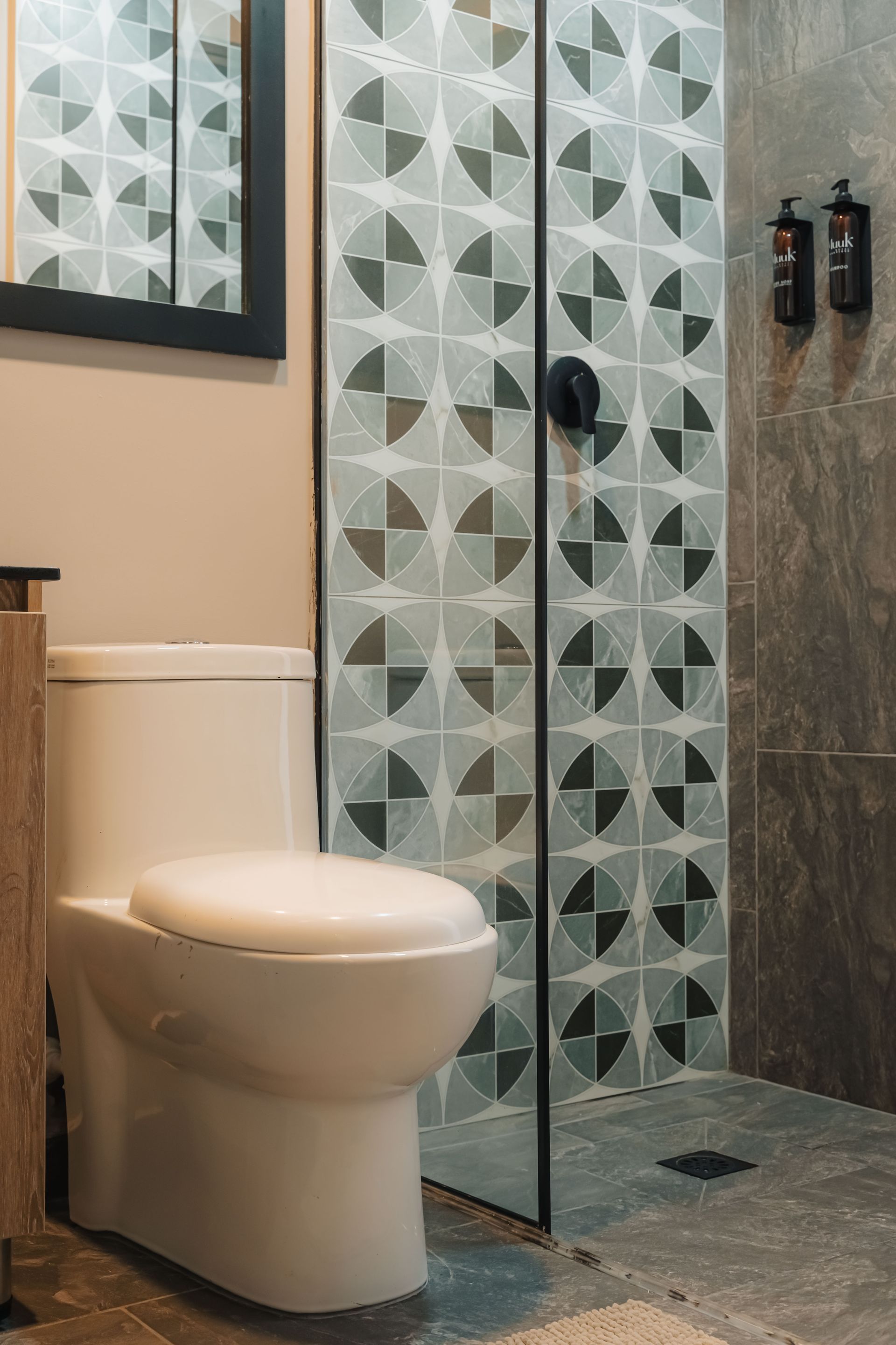 Baño con inodoro blanco, ducha de cristal con azulejos estampados y paredes de color gris oscuro.