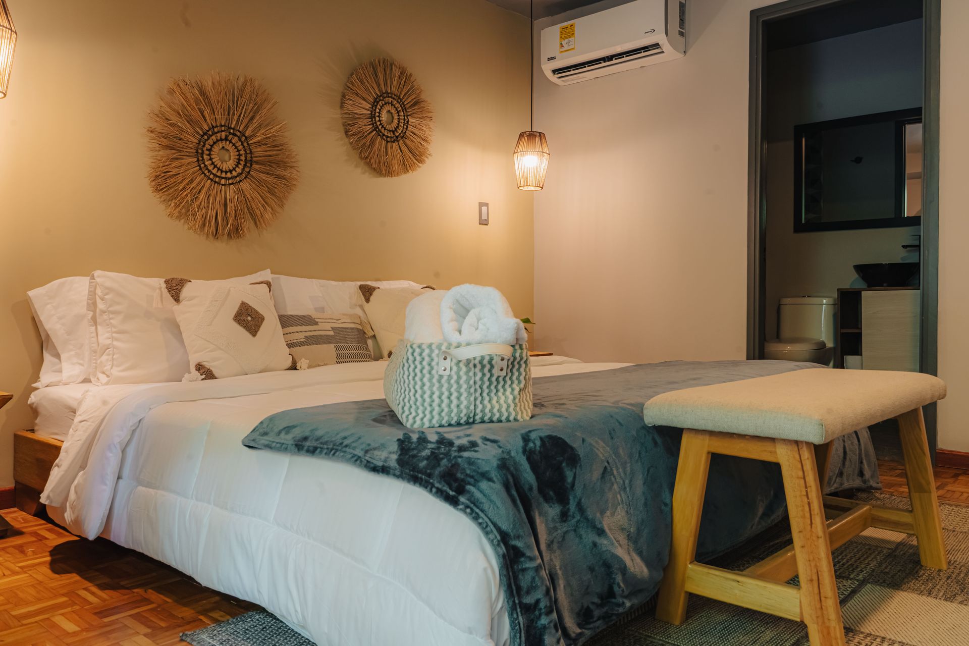 Dormitorio con cama, almohadas, manta, taburete, tapices tejidos y puerta de baño.