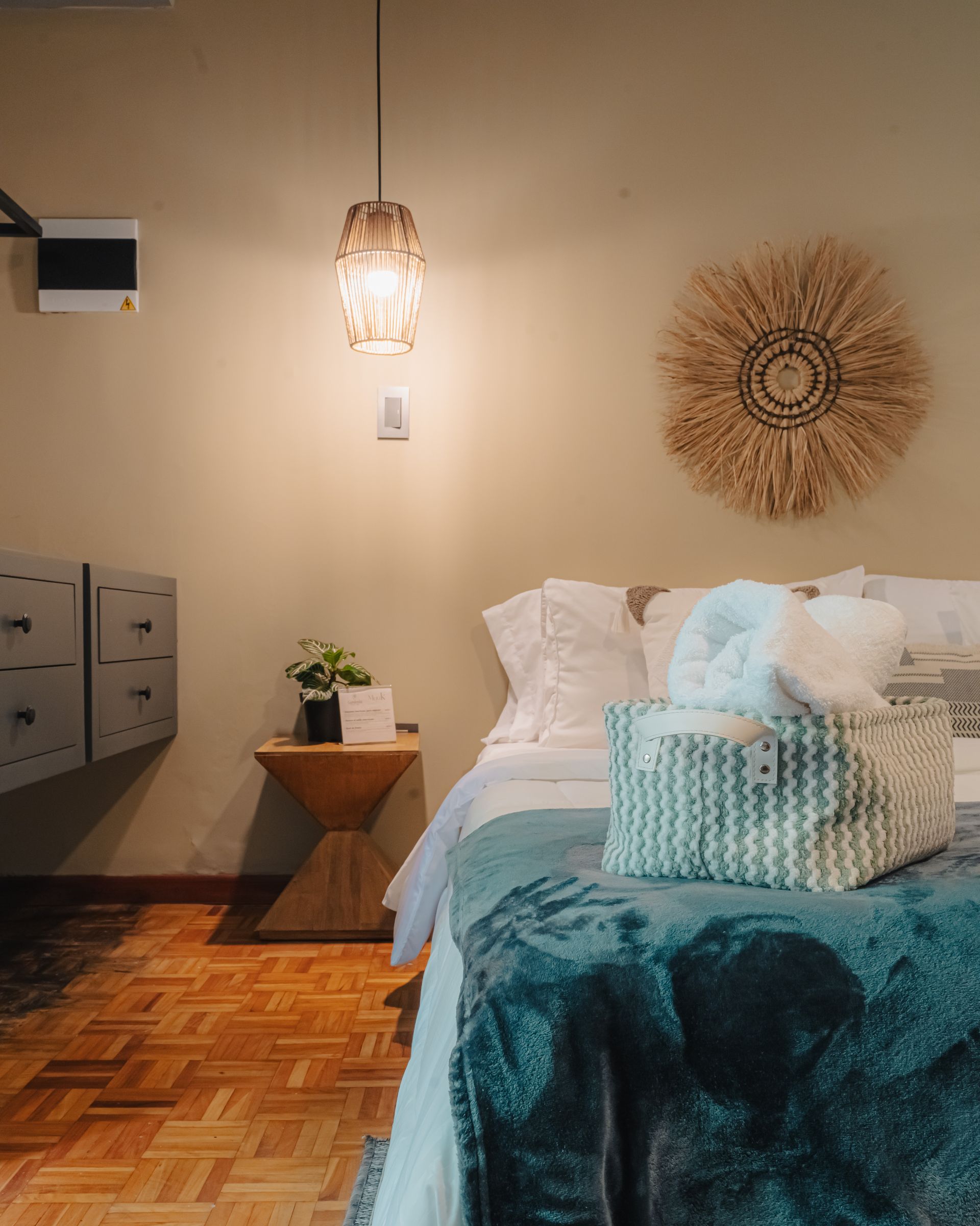 Dormitorio con cama, mesita de noche, piso de madera estampado, lámpara tejida y arte de pared.