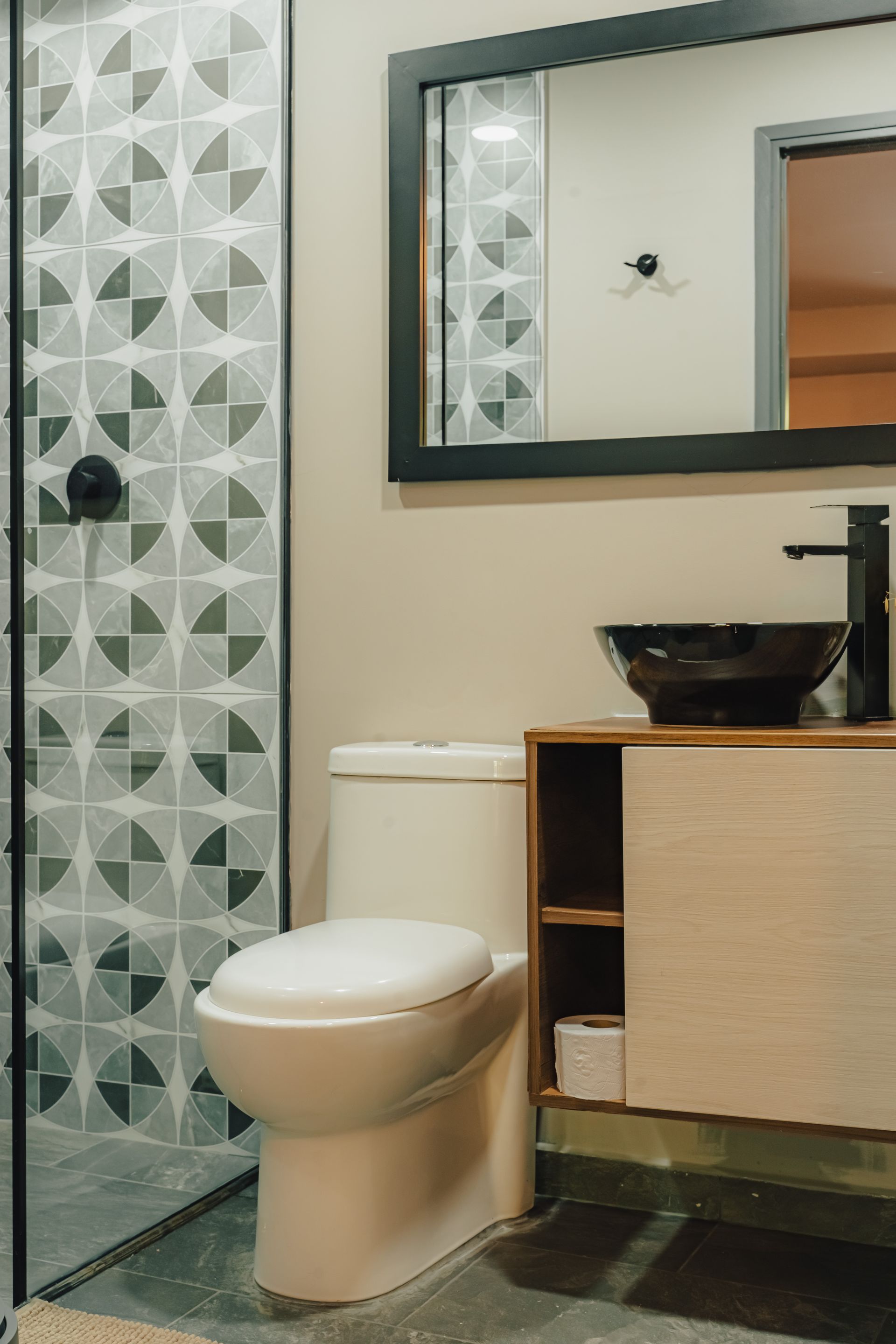 Baño con ducha con azulejos geométricos, inodoro, espejo con marco negro y tocador de madera con lavabo negro.