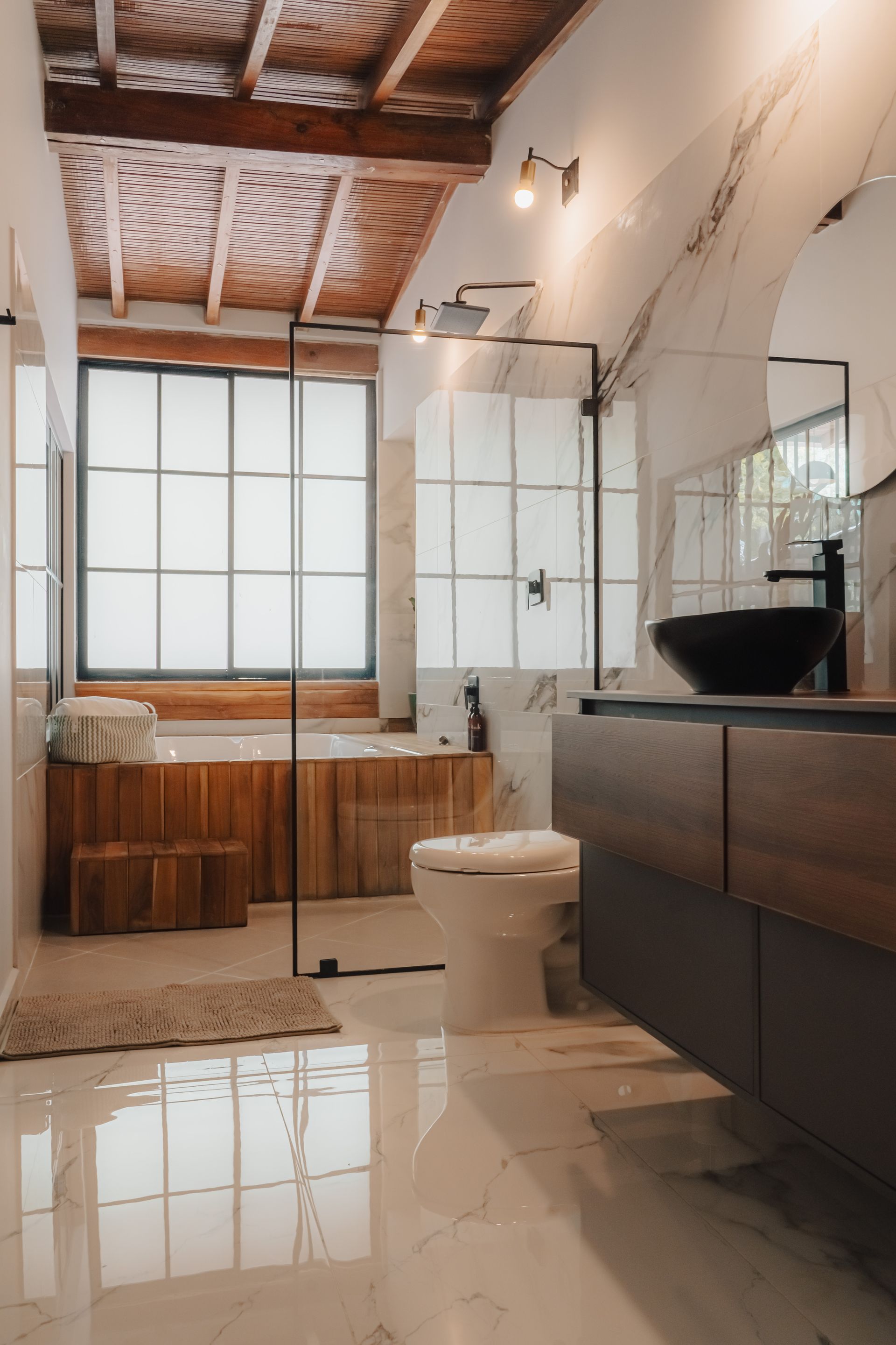 Baño moderno con detalles en madera, grandes ventanales, ducha de cristal, lavabo negro y suelo de mármol blanco.