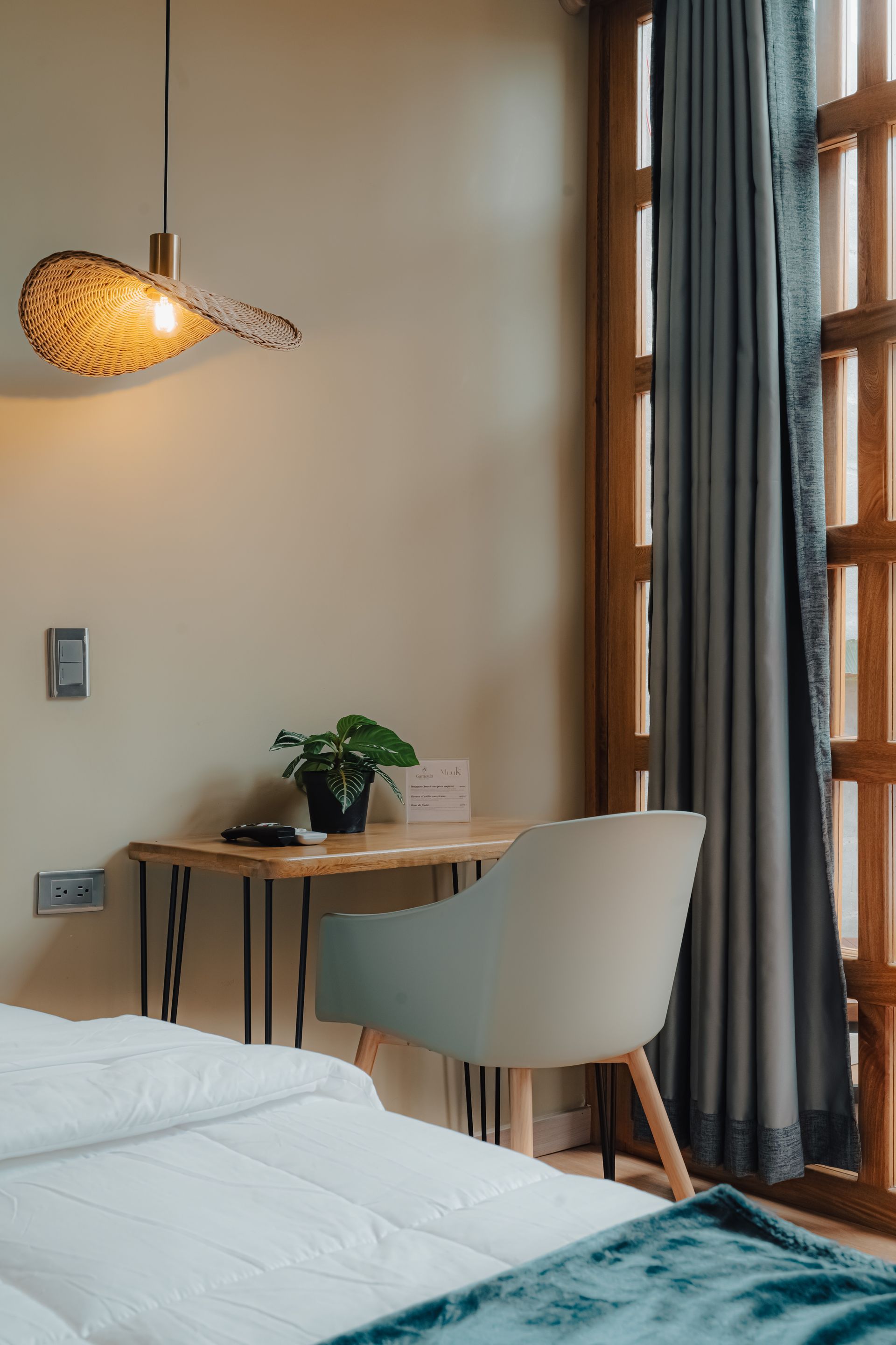 Dormitorio con pequeño escritorio, silla, ventana, lámpara colgante y cama parcialmente visible con manta azul.