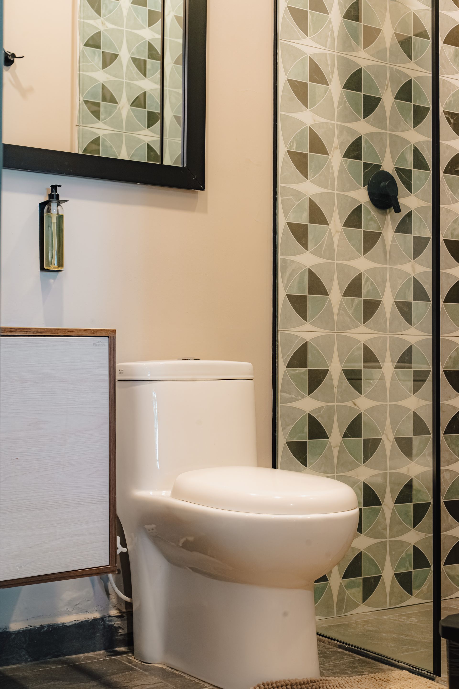 Inodoro blanco en un baño con azulejos de pared estampados, espejo con marco negro y ducha.